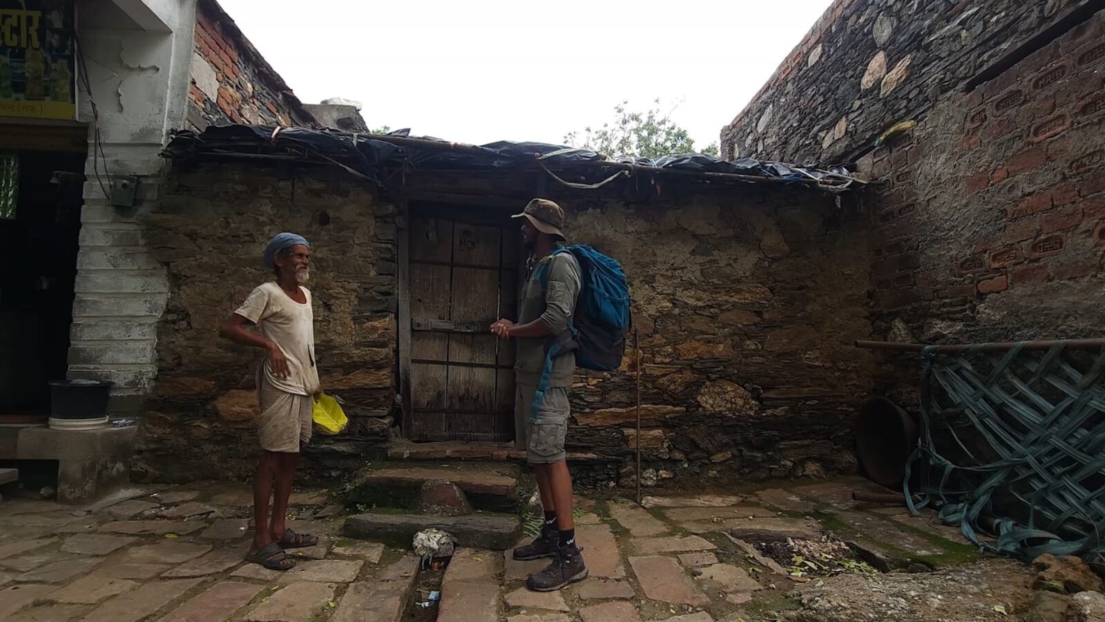 The filmmaker, with a trekking backpack, talking with Bhairaram Ji outside his stone home in Pasun village