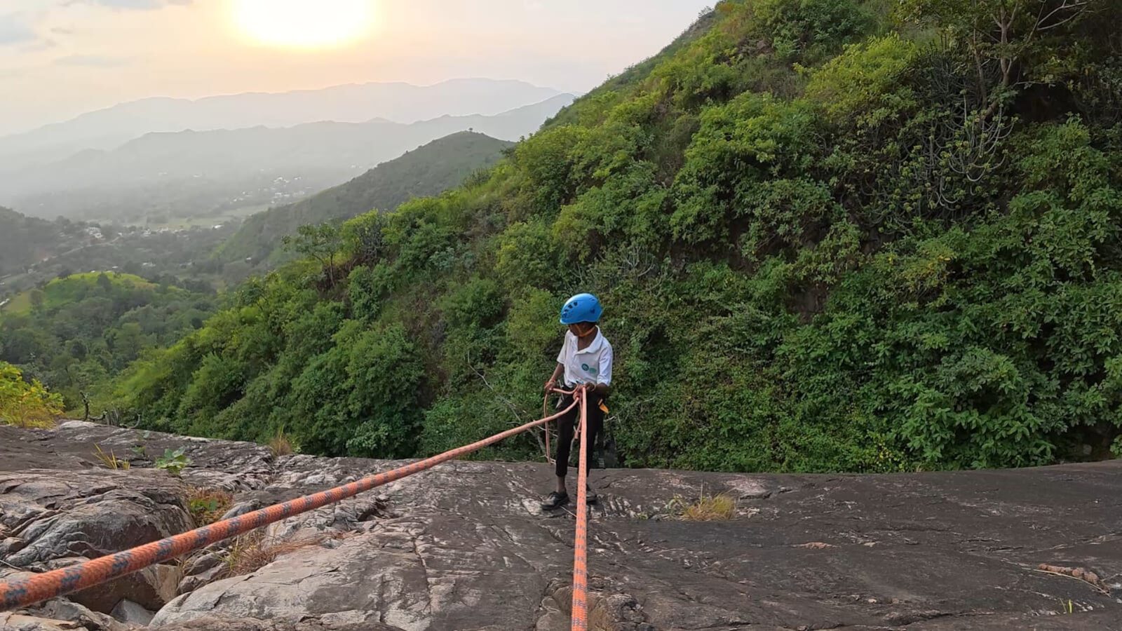 A young boy rappelling down a granite wall at sunset in the Aravalis