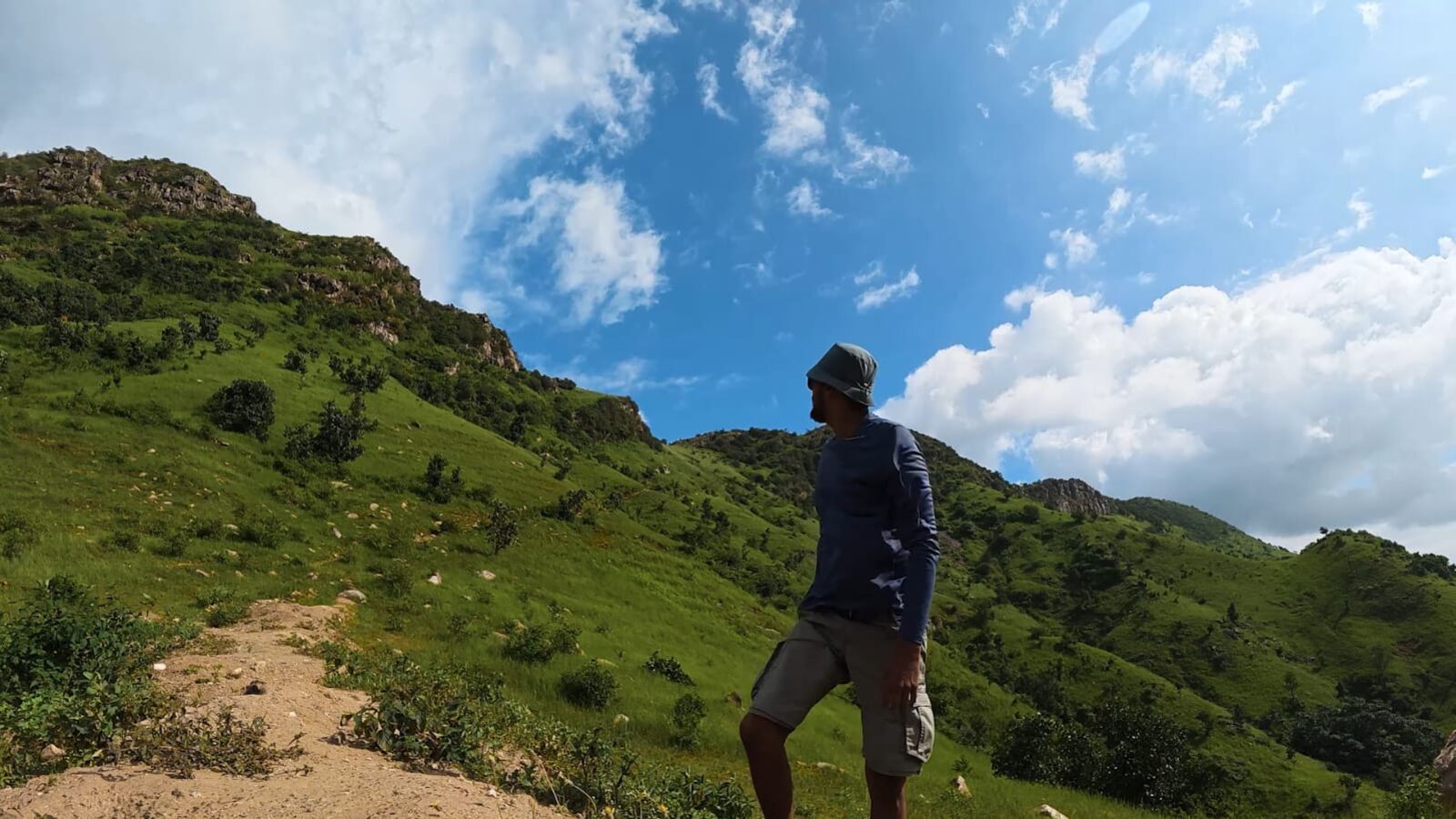 A trekker climbing the green approach toward the Shiva Valley ridgeline under a bright sky